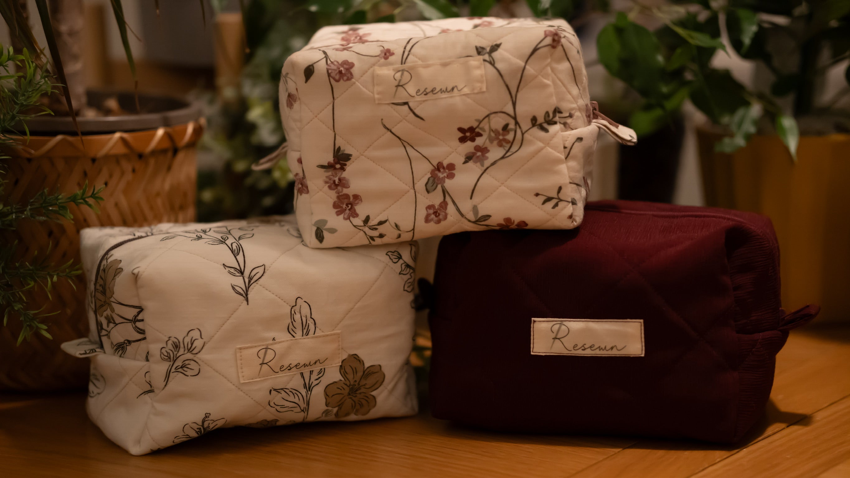 A stack of boxy makeup bags on a wooden surface with plants in the background