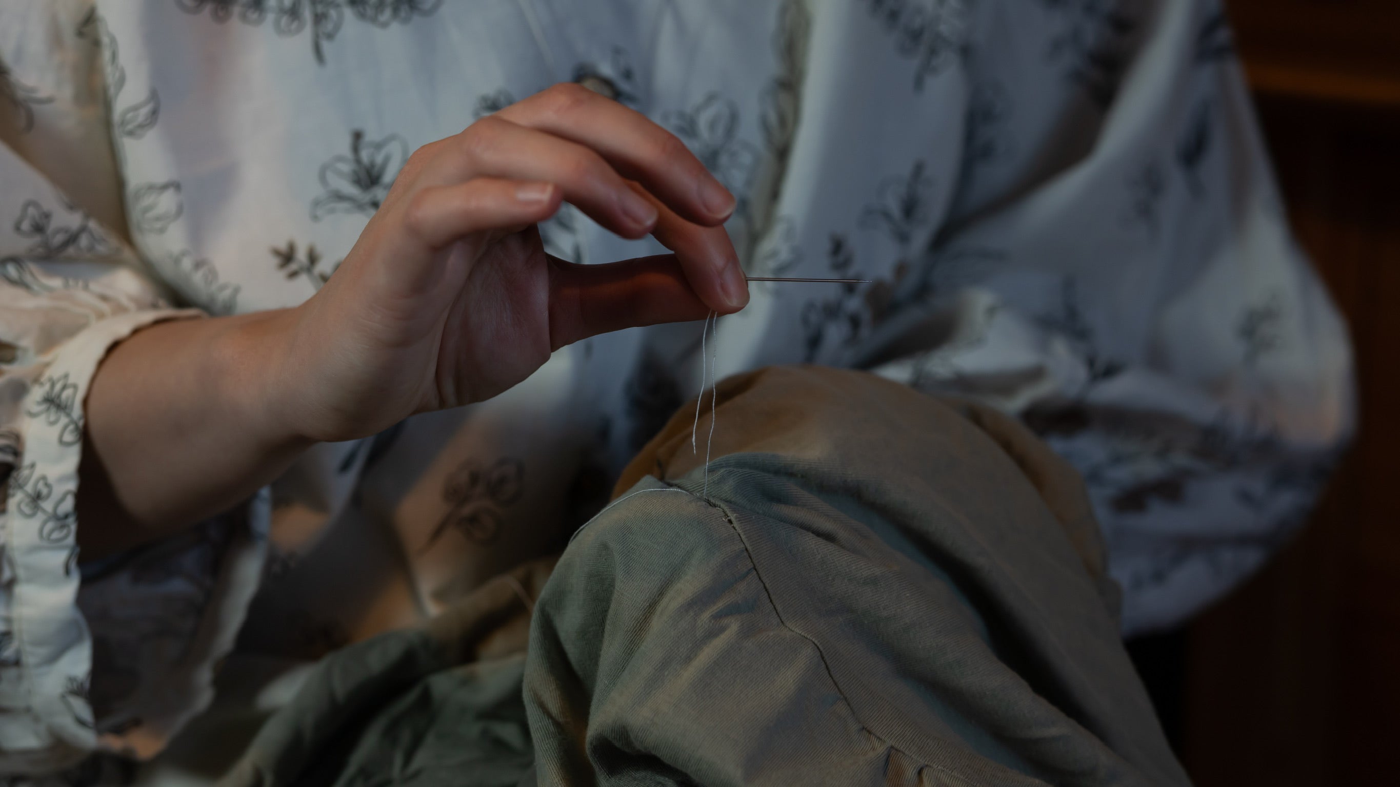 Person sewing with a needle and thread, wearing a floral-patterned garment.