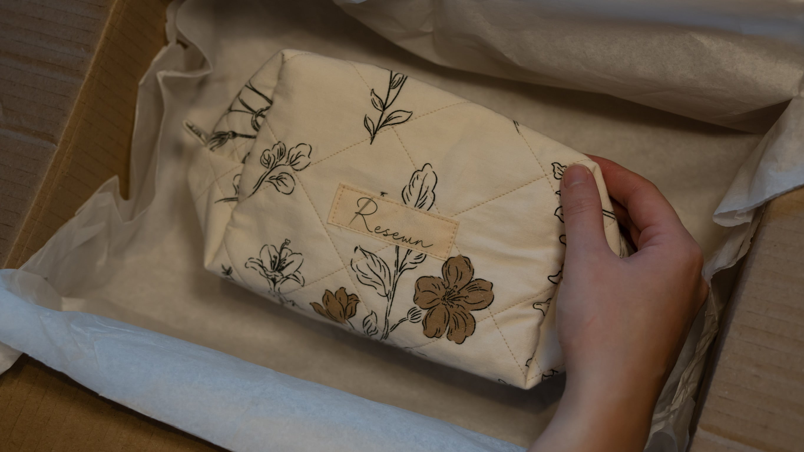 Hand holding a wrapped product with floral designs and a visible brand name inside a box.