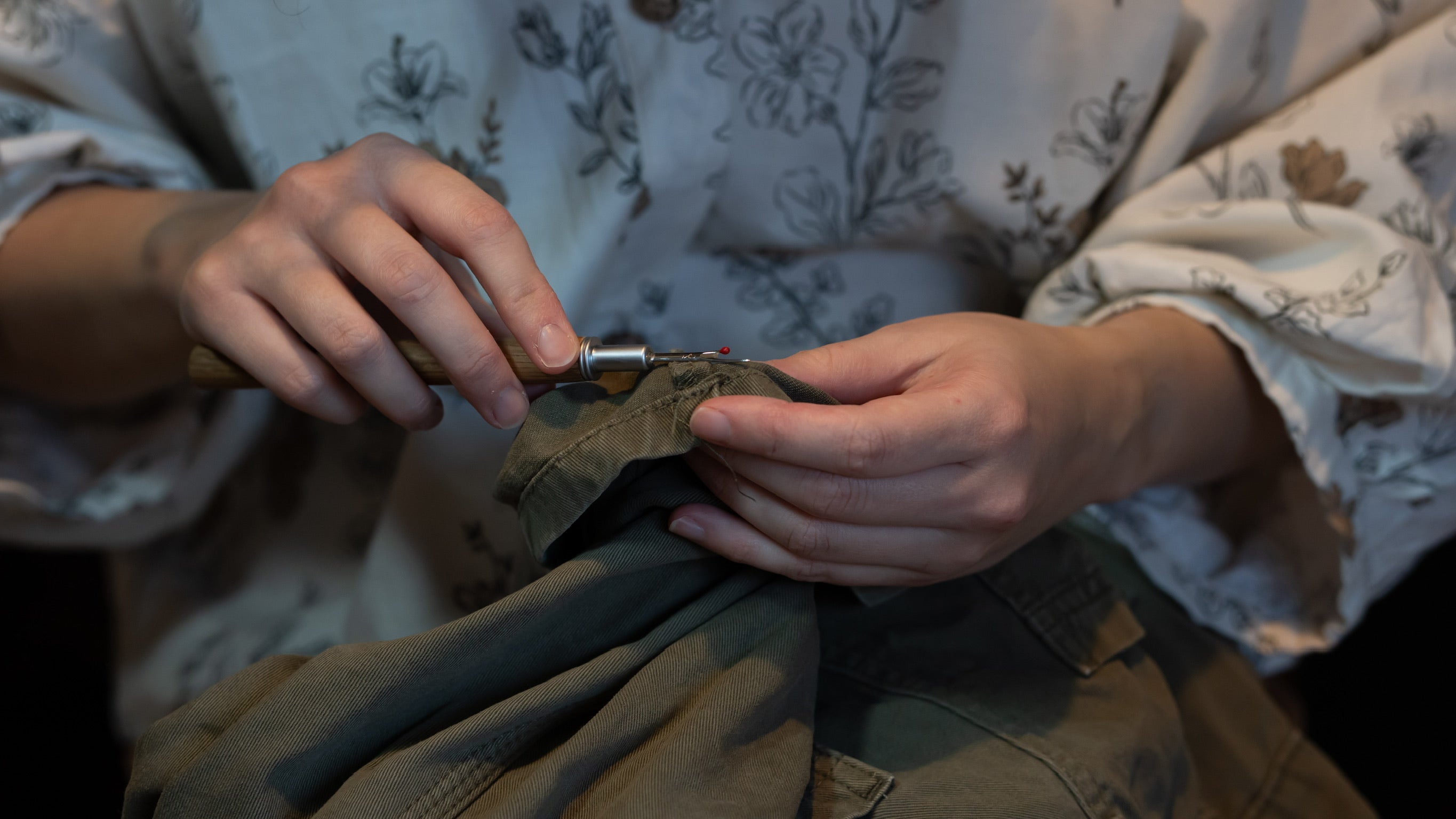Person unpicking the hem of some trousers with a tool, wearing a floral shirt.