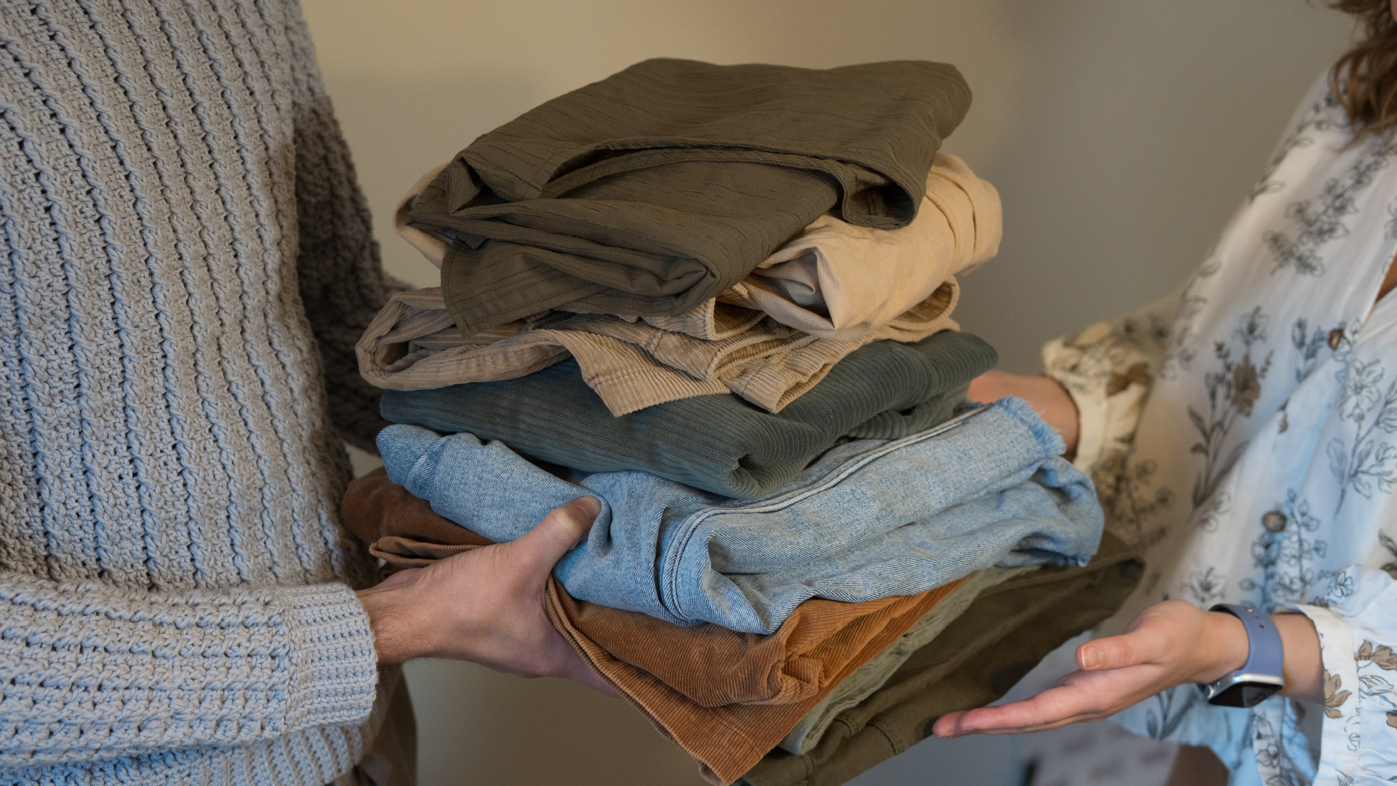 Person holding a stack of folded clothes  passing to another person against a neutral background