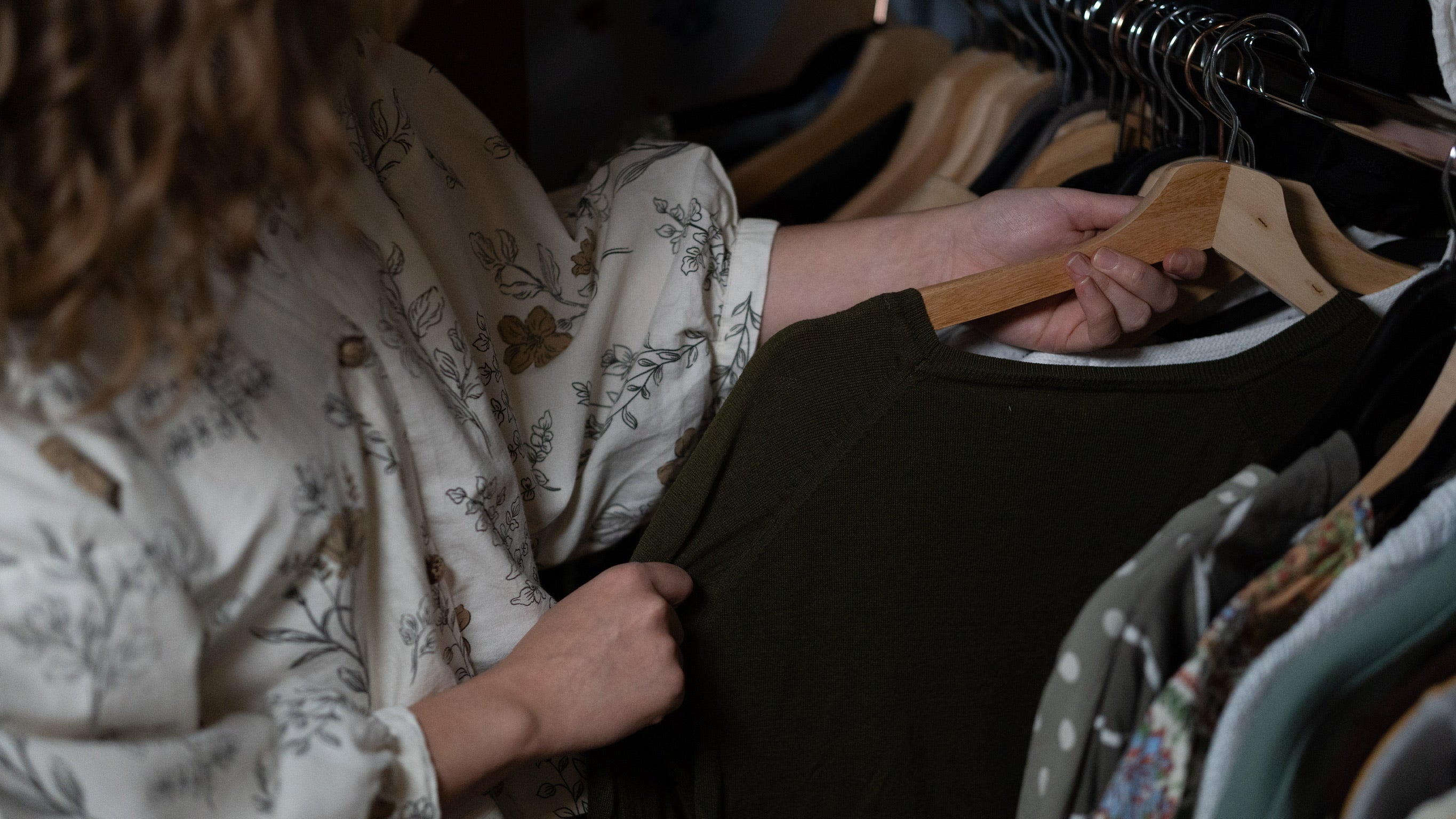 Person holding a green garment in front of a rack with various clothing items in a wardrobe