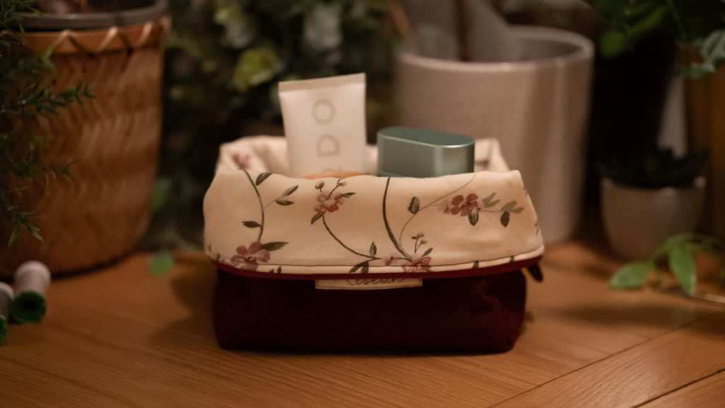 Small floral-patterned boxy bag opened with cosmetic items on a wooden surface, surrounded by potted plants.