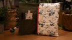 Floral-patterned book sleeve opened to show pockets on a wooden surface with plants in the background