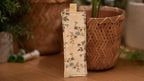 Floral-patterned bookmark on a wooden surface with plants in the background