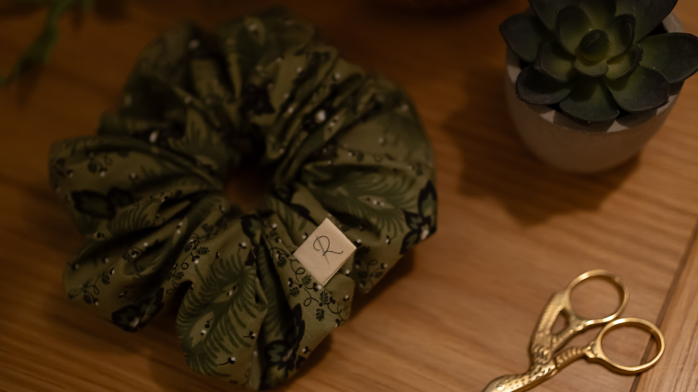Green scrunchie with a brand tag on a wooden surface with gold scissors and a small plant.