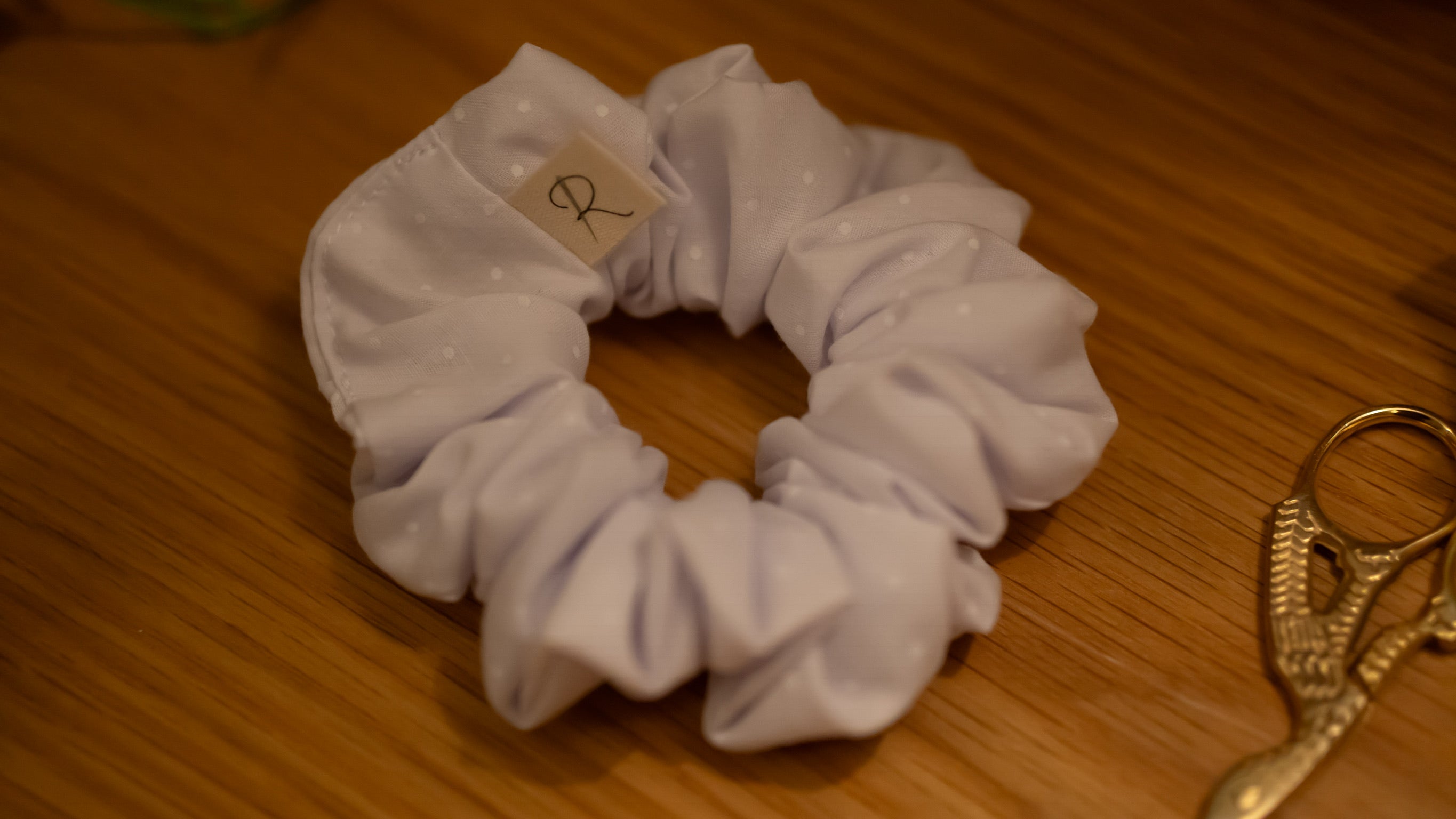 Lilac polka dot scrunchie with a label on a wooden surface next to a gold scissors 