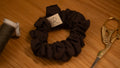 Brown scrunchie with a small label on a wooden surface next to gold scissors.